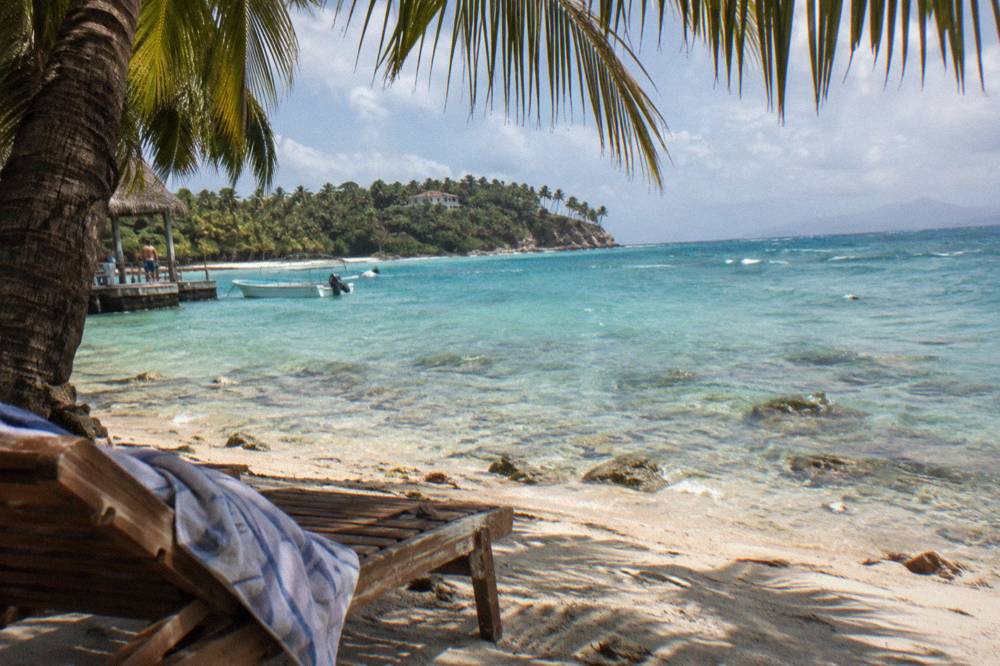 An empty palm-shaded beach with turquoise shallows and a wooden sun lounger