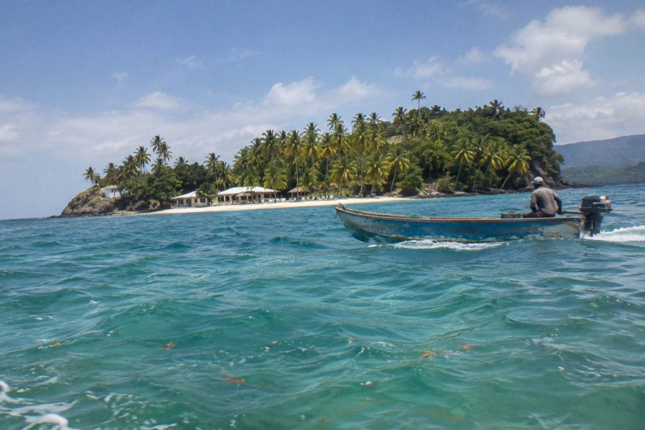 A small palm-covered island with a pale sand beach seen across calm turquoise water