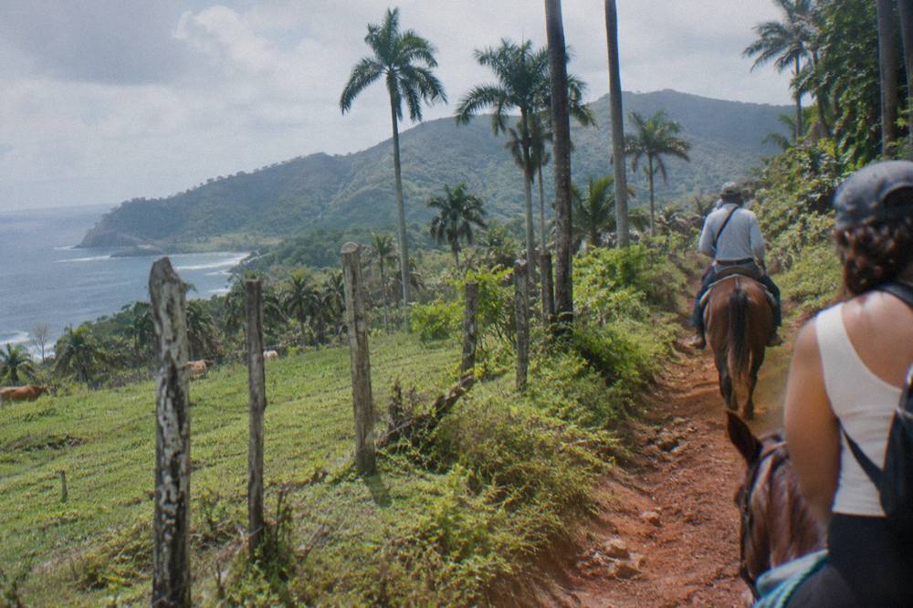 Riders on horseback following a guide along a narrow trail through green farmland toward the hills