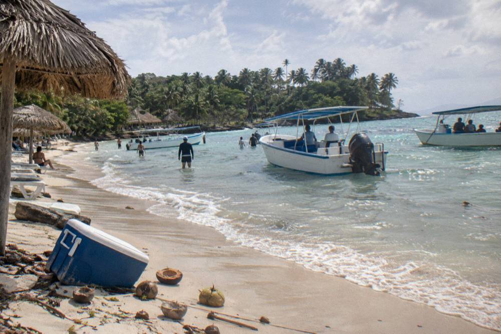 Tour boats moored in shallow water beside a small island beach with sun umbrellas