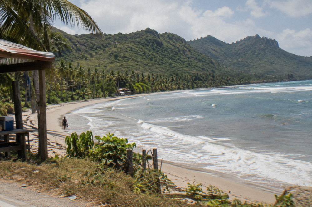 A quiet curved beach backed by palms and green hills at the end of a peninsula road