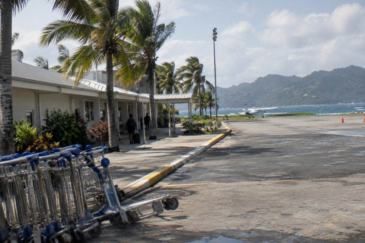 Small regional airport terminal building beside a palm-lined road on a green peninsula under a clear sky
