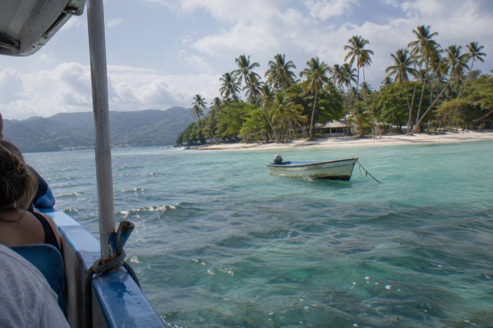 A small island with palm trees and pale sand seen across calm turquoise water.