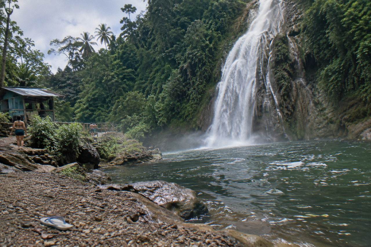 A tall waterfall dropping into a forested pool among green hills