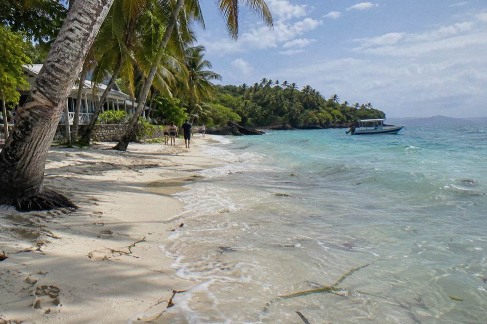 A palm-lined white sand beach with turquoise shallows and a small boat at anchor